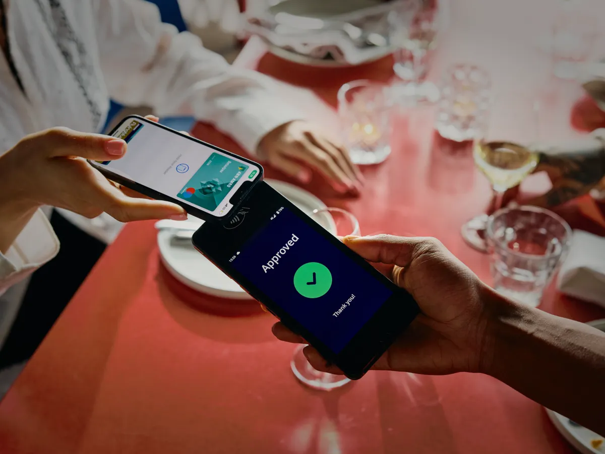 A card transaction between a phone and a terminal is in progress. The terminal displays a green checkmark and the text 'Approved'.