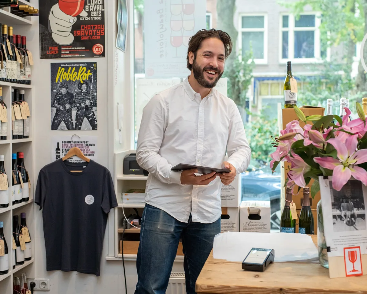Man holding a tablet and smiling to the camera while standing inside a wine shop.